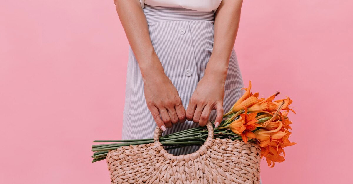 lady-poses-with-knitted-bag-flowers-pink-background-woman-blue-light-skirt-holds-straw-handbag-with-orange-beautiful-bouquet